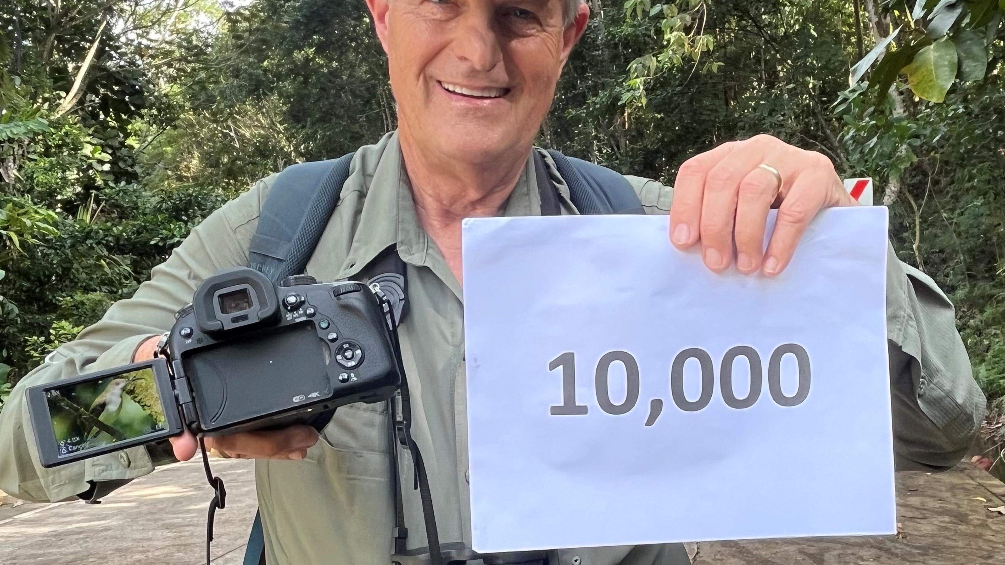 HANDOUT - 09.04.2024, Philippinen, -: Vogelbeobachter Peter Kaestner hält seine Kamera und ein Schild mit der Aufschrift «10.000». Nummer 10 000 versteckte sich in einem Dickicht aus Helikonien auf der philippinischen Insel Mindanao. Eigentlich seien er und sein Helfer Zardo Goring an diesem Nachmittag im Februar gerade auf der Suche nach einer Spechtart gewesen, sagt Peter Kaestner. Doch dann hörten sie ein Geräusch aus dem Helikoniendickicht - und ein grün-orangen Nektarvogel (Arachnothera flammifera) tauchte auf. (zu dpa "Vogel-Beobachtung extrem: US-Amerikaner sichtet 10 000 Arten") Foto: William Suerte/Peter Kaestner/dpa - ACHTUNG: Nur zur redaktionellen Verwendung im Zusammenhang mit der aktuellen Berichterstattung und nur mit vollständiger Nennung des vorstehenden Credits +++ dpa-Bildfunk +++